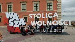 Na tle biało-czerwonego napisi Warszawa stolica wolności 1918-2018 stoi grupa uczestników wycieczki do stolicy wraz z opiekunem.