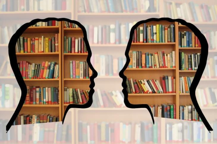 Zdjęcie przedstawia czarne zarysy dwóch głów ludzi na tle półek bibliotecznych pełnych kolorowych książek