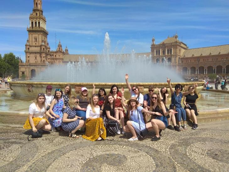 Grupa kolorowo ubranych uczniów wraz z opiekunką na tle dużej fontanny oraz pałacu.