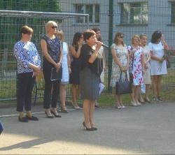 Zdjęcie przedstawia panią Dyrektor przemawiającą podczas uroczystości zakończenia roku szkolnego. Za nią stoi grupa nauczycieli.
