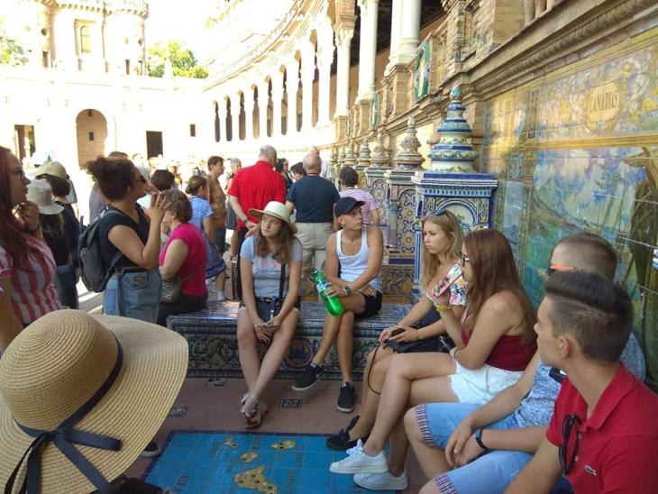 Grupa roześmianych geodetów ubranych kolorowo odpoczywa podczas zwiedzania Sewilli na tle budynku pokrytego piękną mozaiką..
