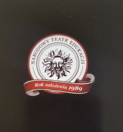Na czarnym tle logo Narodowego Teatru Edukacji - maska teatralna na tle słońca.