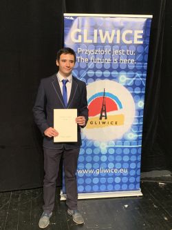 Na zdjęciu Laureat Gliwickiego Lauru Nauki w galowym stroju na tle baneru reklamującego Gliwice