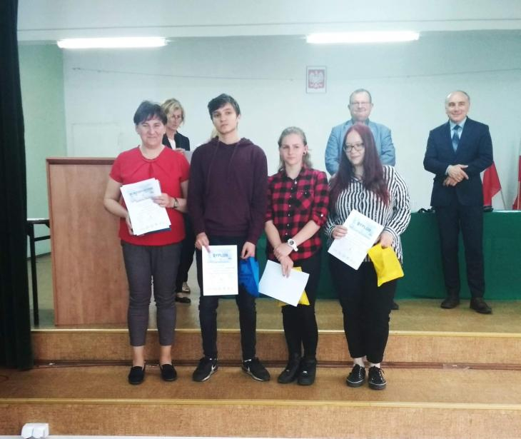 Na zdjęciu znajduje się troje uczniów trzymających dyplomy, ich opiekuna, a w tle widać organizatorów.
