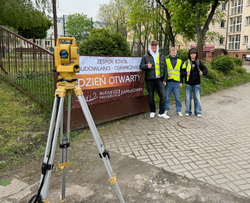 Dzień Otwarty - uczniowie stoją przy bramie wejściowej, na której wisi baner z napisem Dzień Otwarty ZSB-C, przed nimi stoi teodolit