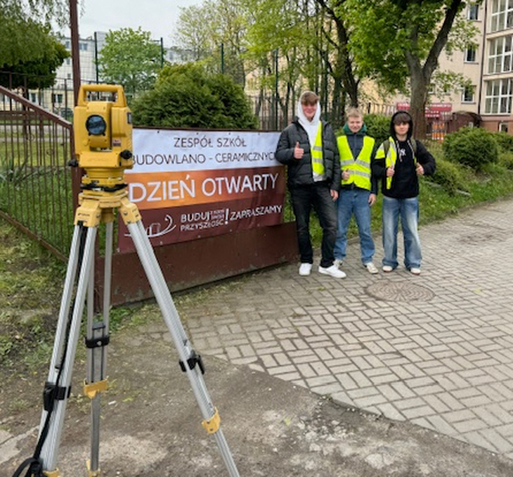 Dzień Otwarty - uczniowie stoją przy bramie wejściowej, na której wisi baner z napisem Dzień Otwarty ZSB-C, przed nimi stoi teodolit