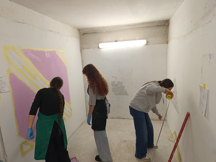 Na zdjęciu widać trzy osoby pracujące w pomieszczeniu o białych, częściowo nieotynkowanych ścianach. Dwie z nich zajmują się malowaniem fragmentu ściany, który został zabezpieczony taśmą malarską i wypełniony kolorem fioletowym. Trzecia osoba mierzy ścianę za pomocą miarki, prawdopodobnie przygotowując miejsce do dalszych prac. Wszystkie osoby mają na sobie robocze ubrania, a dwie z nich noszą niebieskie rękawiczki ochronne. Pomieszczenie oświetla pojedyncza świetlówka zamontowana na suficie. Atmosfera wskazuje na zajęcia praktyczne lub warsztaty z zakresu renowacji lub dekoracji wnętrz.