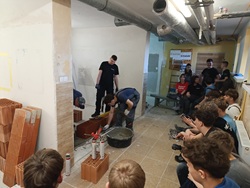 Kilku uczestników stoi przy ścianie z pustaków, gdzie jeden z nich rozprowadza zaprawę z wiadra, a obok ustawione są ceramiczne pustaki. Pozostałe osoby siedzą na ławkach i z zainteresowaniem przyglądają się demonstracji. W tle znajdują się materiały szkoleniowe i plansze, a całość wygląda jak zajęcia praktyczne prowadzone przez specjalistę w ramach szkolenia budowlanego.