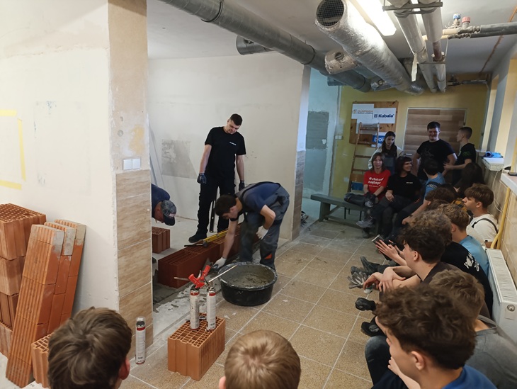 Kilku uczestników stoi przy ścianie z pustaków, gdzie jeden z nich rozprowadza zaprawę z wiadra, a obok ustawione są ceramiczne pustaki. Pozostałe osoby siedzą na ławkach i z zainteresowaniem przyglądają się demonstracji. W tle znajdują się materiały szkoleniowe i plansze, a całość wygląda jak zajęcia praktyczne prowadzone przez specjalistę w ramach szkolenia budowlanego.