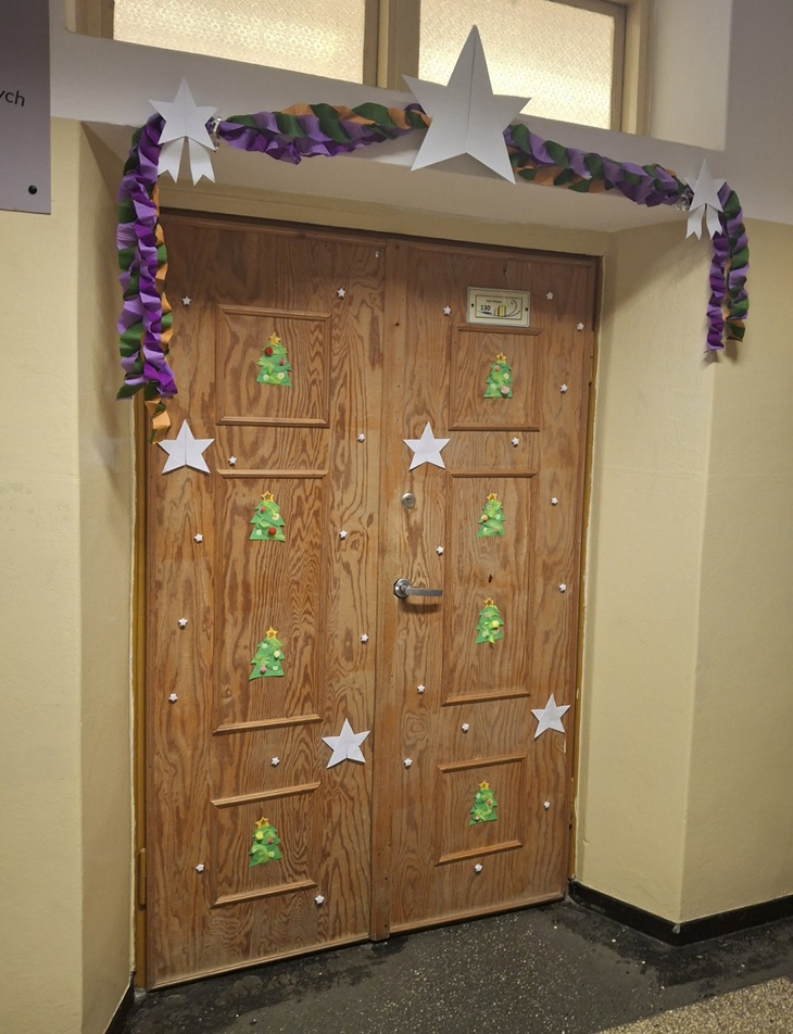 Na zdjęciu widać świątecznie udekorowane drzwi szkolnej sali. Drewniane drzwi ozdobione są papierowymi choinkami oraz małymi białymi gwiazdkami, rozmieszczonymi równomiernie na całej powierzchni.  Nad drzwiami zawieszona jest kolorowa, skręcana girlanda w odcieniach zielonego, fioletowego i pomarańczowego. Girlandę podtrzymują duże, białe papierowe gwiazdy, które dodają dekoracji zimowego i odświętnego charakteru. Całość tworzy radosną, świąteczną atmosferę i wygląda jak element konkursu na najładniej udekorowane wejście do sali.