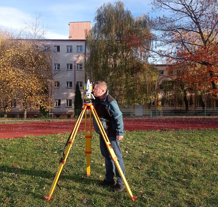 Na zdjęciu widać mężczyznę pracującego w terenie z niwelatorem lub innym przyrządem geodezyjnym ustawionym na statywie. Stoi na trawiastej polanie, a w tle znajdują się budynki mieszkalne oraz drzewa w jesiennych barwach.