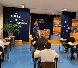 Na zdjęciu widać salę szkolną podczas konkursu „Złota Kielnia” w Gliwicach 2025. Uczniowie siedzą w ławkach, a prowadząca stoi z przodu przy stanowisku z elementami murarskimi, obok banerów szkoły budowlanej.