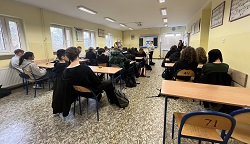 Na zdjęciu widać klasę szkolną, w której uczniowie siedzą przy ławkach i uczestniczą w zajęciach. W tle znajduje się tablica, a nauczyciel lub prowadzący stoi przy niej i mówi do grupy. Uczniowie wydają się skupieni na zajęciach. 