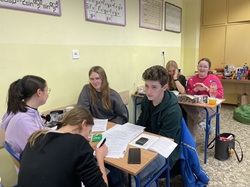 To zdjęcie przedstawia grupę młodych ludzi siedzących w sali lekcyjnej. Widać, że pracują nad zadaniami matematycznymi – na stołach znajdują się arkusze, kalkulatory i przybory do pisania. Niektórzy uczestnicy są skupieni, inni się uśmiechają, co sugeruje, że mimo wysiłku atmosfera jest przyjazna. W tle widać tablice z wzorami matematycznymi oraz przekąski i napoje, co może wskazywać na długą sesję nauki lub konkurs matematyczny. 