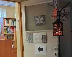 Wejście do szkolnej biblioteki udekorowane na Halloween. Po prawej stronie wisi czarna latarnia z pomarańczowym napisem „Boo!”, ozdobiona motywami duchów, obok znajdują się pajęczyny. Na drzwiach wiszą ilustracje o tematyce literackiej, informacja o godzinach otwarcia biblioteki oraz strzałka wskazująca wejście. Po lewej stronie widać regały z książkami i drewnianą ladę.