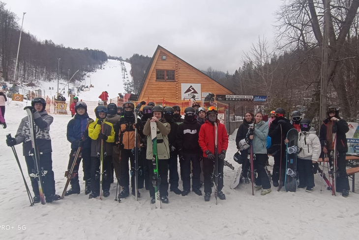 Grupa narciarzy i snowboardzistów pozuje do zdjęcia u podnóża stoku narciarskiego w zimowej scenerii.