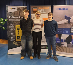 Na zdjęciu widać trzy osoby stojące obok siebie na tle banerów promocyjnych związanych z olimpiadą „Buduj z Pasją”. Po bokach znajdują się reklamy firm związanych z branżą budowlaną. Osoby pozują do zdjęcia w sali szkolnej