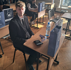 uczeń siedzi przy stoliku, na którym stoi butelka wody, dwie torby papierowe od sponsora oraz kalkulator i długopis