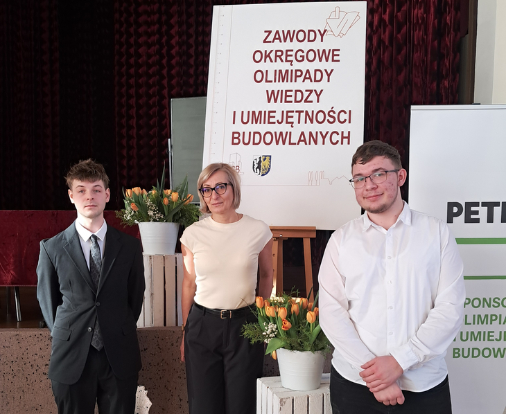 Na zdjęciu widać trzy osoby stojące przed tablicą z napisem „Zawody Okręgowe Olimpiady Wiedzy i Umiejętności Budowlanych”. Po bokach ustawione są dekoracje z kwiatami w donicach. Dwie osoby stoją po lewej i prawej stronie tablicy, a jedna znajduje się pośrodku. W tle widać czerwone zasłony oraz baner sponsora wydarzenia.