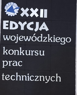 Na zdjęciu widać tablicę informacyjną ustawioną na sztaludze z napisem „XXII edycja wojewódzkiego konkursu prac technicznych”. Obok znajduje się stolik oraz dekoracyjna roślina