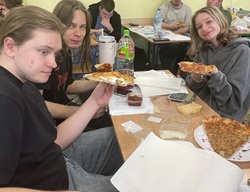 Grupa uczniów podczas maratonu matematycznego robi przerwę na pizzę 🍕. Siedzą przy ławce z arkuszami i kalkulatorem, uśmiechnięci i wyraźnie zadowoleni z chwili odpoczynku po intensywnej pracy