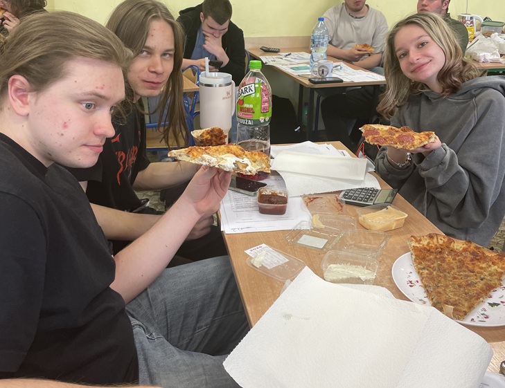 Grupa uczniów podczas maratonu matematycznego robi przerwę na pizzę 🍕. Siedzą przy ławce z arkuszami i kalkulatorem, uśmiechnięci i wyraźnie zadowoleni z chwili odpoczynku po intensywnej pracy