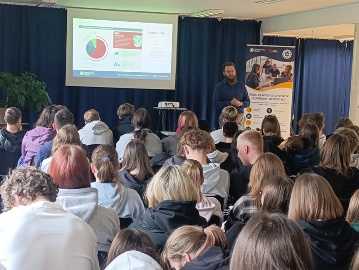 Na zdjęciu widać uczniów zgromadzonych na sali podczas prelekcji o cyberbezpieczeństwie. Prowadzący stoi przed ekranem z prezentacją, omawiając zagadnienia związane z zagrożeniami w sieci, a uczestnicy uważnie słuchają.