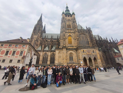 Na zdjęciu widać grupę uczniów stojących przed Katedra św. Wita w Pradze. W tle dominuje imponująca gotycka budowla z wysokimi wieżami, a uczniowie pozują do wspólnego zdjęcia podczas wycieczki.