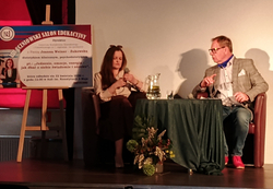 Na zdjęciu widać spotkanie edukacyjne odbywające się na scenie. Dwie osoby siedzą w fotelach przy stoliku, rozmawiając – obok stoi dzbanek z wodą i szklanka. Po lewej stronie znajduje się plansza informująca o wydarzeniu „Gliwicki Salon Edukacyjny”, dotyczącym m.in. zdrowia i świadomego stylu życia.