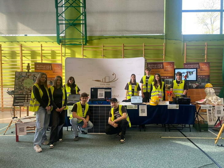 Na zdjęciu widać grupę młodych osób stojących przy stanowisku wystawowym w hali sportowej. Wszyscy mają na sobie odblaskowe kamizelki, a przed nimi znajdują się różne materiały i sprzęt – m.in. panel słoneczny, modele oraz narzędzia związane z budownictwem lub techniką. W tle są banery promujące szkołę oraz sprzęt prezentacyjny. Atmosfera przypomina targi edukacyjne
