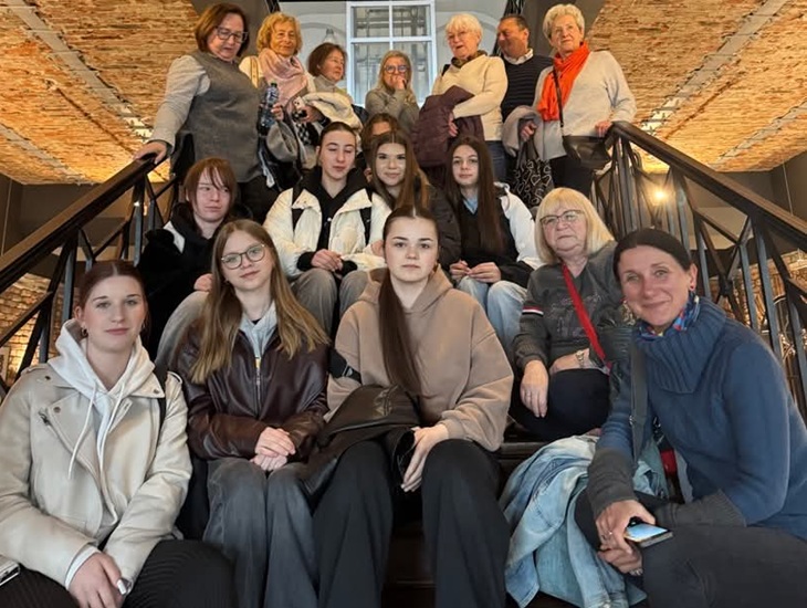 Na zdjęciu widać grupę uczniów wraz z opiekunami siedzących i stojących na schodach w budynku o industrialnym, odnowionym wnętrzu z ceglanego stropu. Wszyscy pozują do wspólnego zdjęcia, tworząc pamiątkową fotografię z wycieczki.