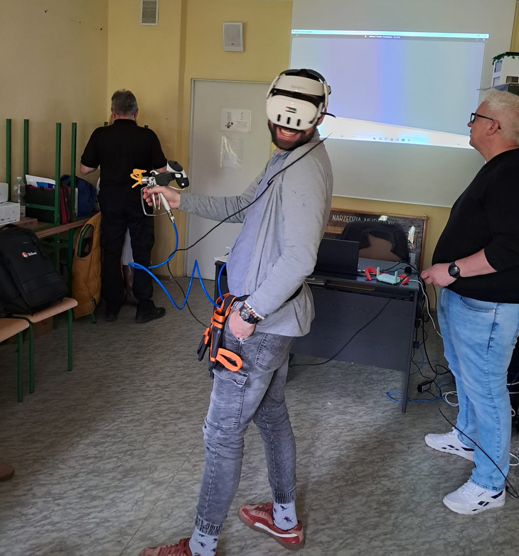 Na zdjęciu nauczyciele uczestniczą w szkoleniu z obsługi symulatora 3D do malowania natryskowego. Jeden z uczestników korzysta z gogli VR oraz pistoletu natryskowego, wykonując ćwiczenie w wirtualnej rzeczywistości.