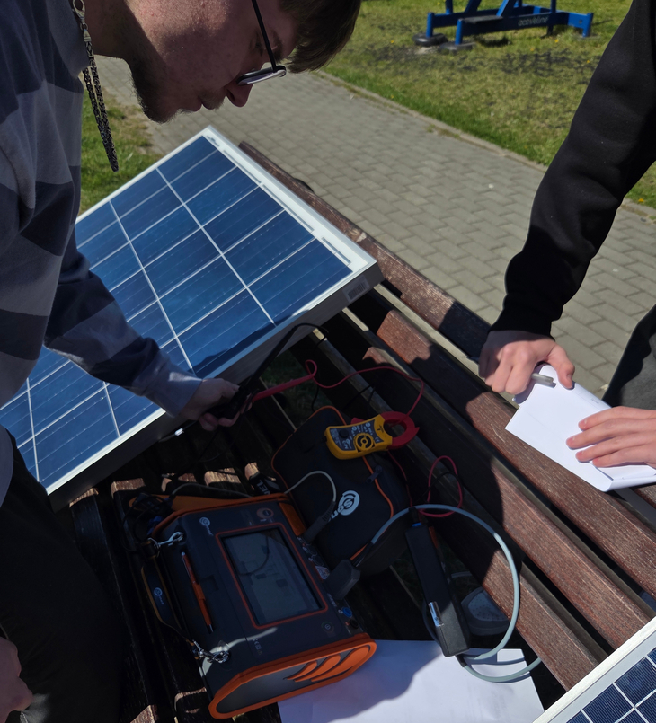 Na zdjęciu uczniowie wykonują pomiary instalacji fotowoltaicznej podczas zajęć praktycznych na świeżym powietrzu. Korzystając ze specjalistycznych mierników i paneli PV, sprawdzają parametry pracy modułów fotowoltaicznych oraz zapisują wyniki pomiarów. Zajęcia pozwalają zdobywać praktyczne umiejętności związane z odnawialnymi źródłami energii.