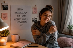 Na zdjęciu widać młodą dziewczynę siedzącą przy biurku w przytulnym, spokojnym pokoju. Ma zamknięte oczy i obejmuje samą siebie, co sugeruje moment wyciszenia, samoakceptacji i troski o własne emocje. Ubrana jest w szarą bluzę z kapturem, a jej swobodna poza tworzy atmosferę bezpieczeństwa i refleksji. W tle znajduje się duży plakat z napisem: „Jestem wystarczająca. Nie muszę niczego nikomu udowadniać”, który stanowi główne przesłanie fotografii. Na ścianie wiszą również małe karteczki motywacyjne, m.in. „Bądź dla siebie dobra” oraz „Twoje tempo jest OK”, podkreślające temat samoakceptacji i łagodności wobec siebie. Pomieszczenie oświetlone jest ciepłym światłem lampki i świecy, co nadaje zdjęciu intymny, spokojny klimat. Na biurku leży notes z narysowanym sercem oraz kubek, które dodatkowo wzmacniają wrażenie codzienności i emocjonalnego komfortu. Całość przedstawia ideę dbania o siebie, odpuszczania presji perfekcjonizmu i budowania poczucia własnej wartości niezależnie od oczekiwań innych.