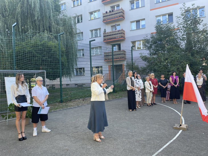 Uroczyste rozpoczęcie roku szkolnego na szkolnym boisku. W centrum dyrektor przemawia do uczniów i grona pedagogicznego, obok stoi polska flaga