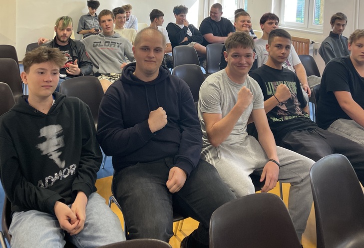Na zdjęciu widać grupę uczniów siedzących w szkolnej sali. W pierwszym rzędzie kilku chłopaków pozuje do zdjęcia, część z nich pokazuje zaciśnięte pięści, jakby podkreślając ducha rywalizacji. W tle siedzą pozostali uczestnicy spotkania, którzy również przyglądają się wydarzeniu.  Zdjęcie oddaje luźną, koleżeńską atmosferę i ducha wspólnej zabawy.