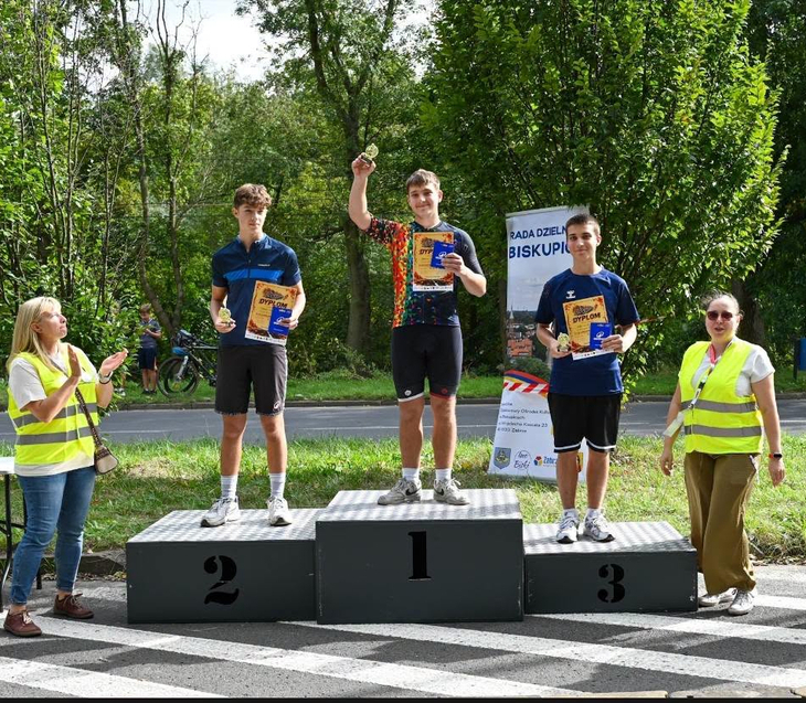 Na zdjęciu widzimy ceremonię wręczenia nagród po wyścigu rowerowym. Trzej zawodnicy stoją na podium, prezentując medale i dyplomy. Na najwyższym stopniu podium znajduje się zwycięzca zawodów. Wokół podium są organizatorzy wydarzenia.