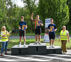 Na zdjęciu widzimy ceremonię wręczenia nagród po wyścigu rowerowym. Trzej zawodnicy stoją na podium, prezentując medale i dyplomy. Na najwyższym stopniu podium znajduje się zwycięzca zawodów. Wokół podium są organizatorzy wydarzenia.