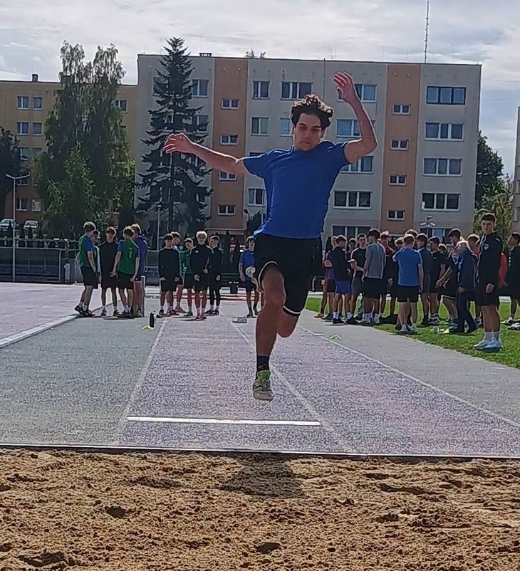 Zawodnik podczas skoku w dal. W tle widać pozostałych uczestników zawodów oraz osoby sędziujące konkurencję. Dynamiczne ujęcie pokazuje moment lotu nad piaskownicą.